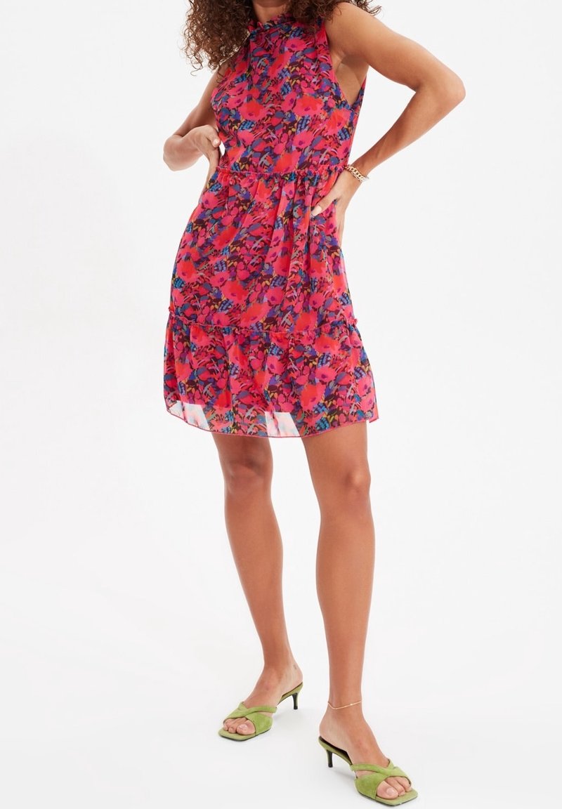 Robe à imprimé floral dans des tons rouges, roses et bleus. Sans manches avec une taille froncée et une jupe à volants. Associée à des sandales ouvertes vertes.
