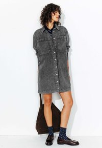 Robe chemise en denim gris avec un devant à boutons, deux poches poitrine et une coupe décontractée, associée à des chaussettes foncées et des mocassins. Sac fourre-tout marron.