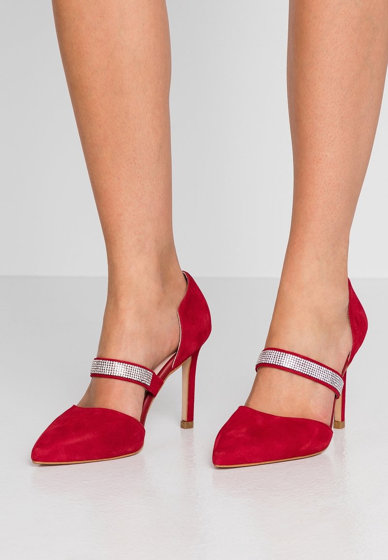 Jambes portant des chaussures à talons hauts en daim rouge avec des bouts pointus et une bride scintillante sur le dessus, debout sur une surface grise lisse.