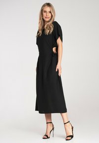 Schwarzes Maxikleid mit weiten Ärmeln, seitlichen Bindungen, die Ausschnitte erzeugen, und einer glatten Textur. Kombiniert mit schwarzen High-Heels.