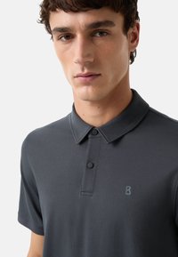 Mørkegrå polo shirt med korte ærmer, krave og to knapper. Har et subtilt struktureret mønster og et lille præget logo på brystet.