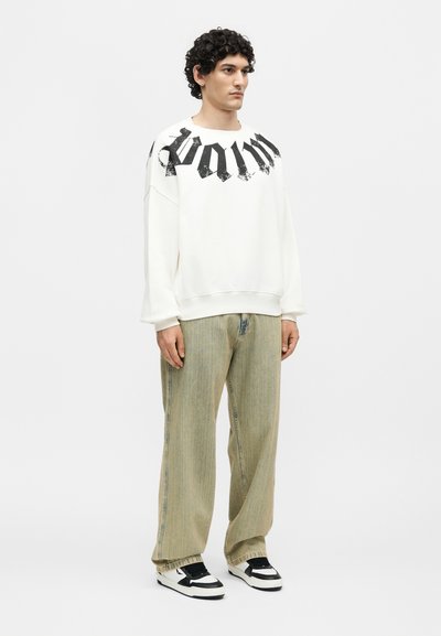 Άνδρας με σγουρά μαλλιά που φοράει λευκό φαρδύ φούτερ με μαύρα γράμματα, άνετο μπεζ τζιν και ασπρόμαυρα αθλητικά παπούτσια, στέκεται ακίνητος.