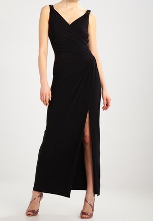 Femme portant une robe de soirée noire sans manches avec une fente latérale et une taille froncée, associée à des sandales métalliques à talons hauts avec des brides.