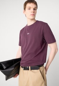 Bordová bavlněná tričko s kulatým výstřihem a malým bílým logem "BOSS" na hrudi, v kombinaci s béžovými kalhotami a černou taškou.