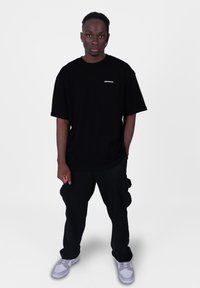 Černé oversized tričko s bílým nápisem "CRYSTAL"; v kombinaci s černými cargo kalhotami a šedými teniskami. Jednoduché pozadí, stojící póza.