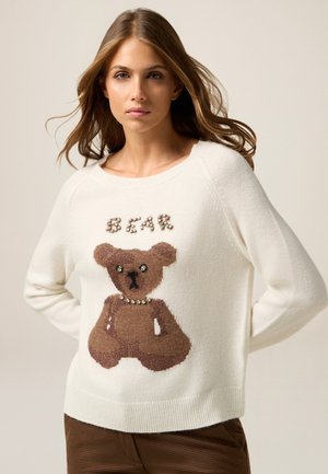 Pull créme avec un design de teddy bear brun, doté d'un fil texturé et d'accents en perles. Encolure ronde simple et poignets côtelés.