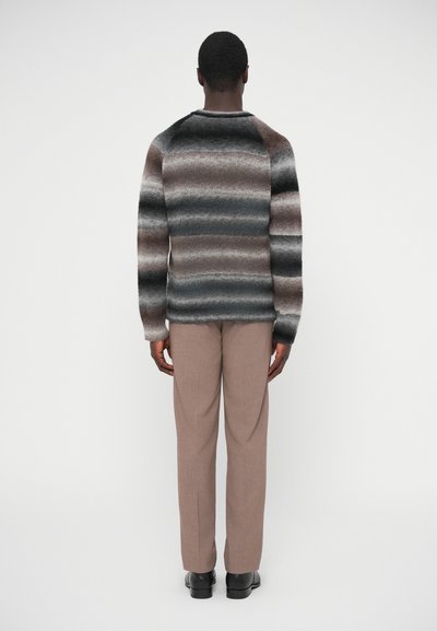 Πλεκτό πουλόβερ με οριζόντιες ρίγες σε σκούρο γκρι, ανοιχτό γκρι και καφέ, στρογγυλή λαιμόκοψη και μακριά μανίκια, συνδυασμένο με μπεζ παντελόνι και μαύρα παπούτσια.