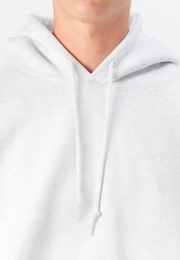 Hoodie cinza claro com uma textura suave, apresentando um bolso canguru, cordões de ajuste e um logótipo bordado no peito.