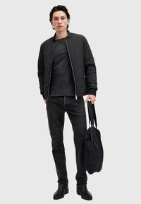 Gequiltete schwarze Bomberjacke, schwarzes Rundhalsoberteil, dunkle Jeans und schwarze Tasche in der Hand. Verfügt über eine glatte Textur und ein einfaches Design.