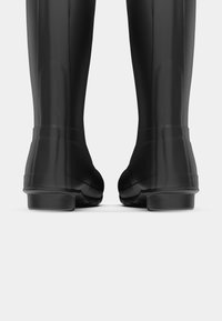 Bottes en caoutchouc noires, imperméables, avec une surface lisse, un bout arrondi, un talon bas et une semelle texturée pour une meilleure adhérence, vues de l'arrière.