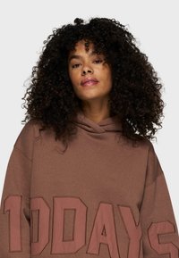 Hoodie marron avec un grand texte "10 JOURS" dans une couleur contrastante. Il possède une capuche avec cordon de serrage, des manches larges et un tissu texturé.