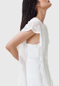 Top bianco con maniche trasparenti ricamate a motivi floreali. Presenta un colletto tondo e una vita elasticizzata per un look aderente.