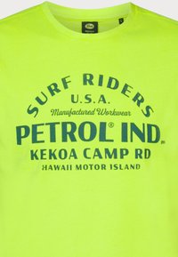 Jasně žluté tričko vyrobené z bavlny, s modrým grafickým potiskem obsahujícím text "Surf Riders U.S.A." a "Petrol Ind." Design s kulatým výstřihem.