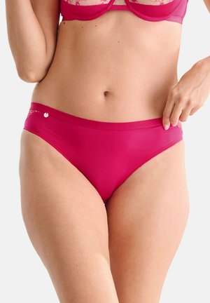Femme ajustant un ensemble de lingerie en dentelle rose, montrant le torse et la main tirant la ceinture sur un fond uni clair.