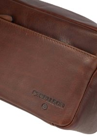 Bolsito de cuero marrón con textura suave, que presenta una costura y el logo "Strellson" en relieve. Forma rectangular compacta.