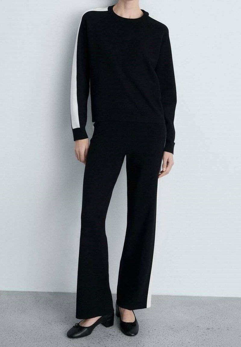 Personne debout portant un pantalon noir et un haut noir à manches longues avec des bandes blanches le long du côté de la manche gauche et du pantalon.