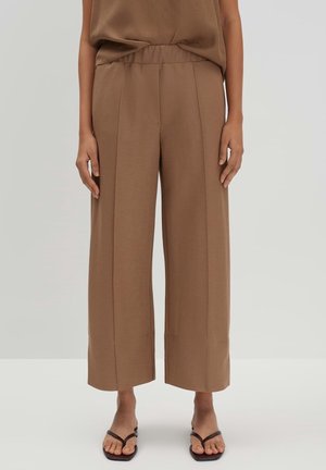 Kobieta ubrana w brązowe spodnie z wysokim stanem i szerokimi nogawkami z przednimi szwami oraz czarne sandały typu japonki, stojąca na jednolitym szarym tle.