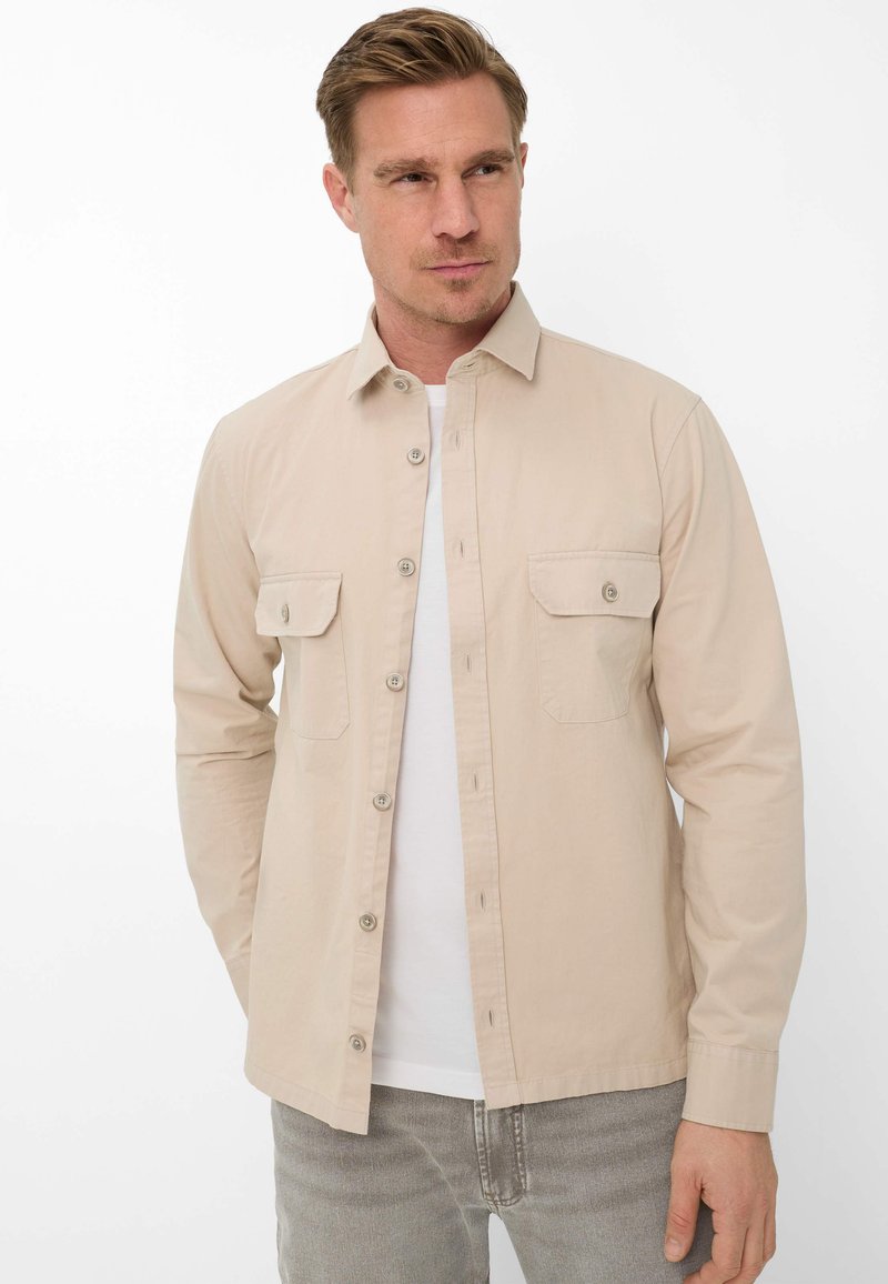 Blonder Mann, der ein beiges Hemd über einem weißen T-Shirt und graue Jeans trägt, mit den Händen in den Taschen steht und zur Seite schaut.