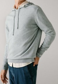 Felpa con cappuccio grigia chiara realizzata in tessuto morbido, con coulisse, polsini e orlo a coste. Indossata sopra una camicia bianca con colletto.