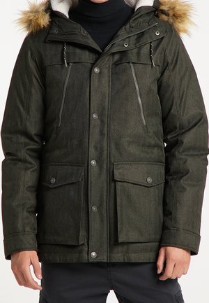 Homme portant une parka d'hiver vert foncé avec capuche bordée de fourrure, plusieurs poches zippées et à boutons, sur un pull à col roulé noir.