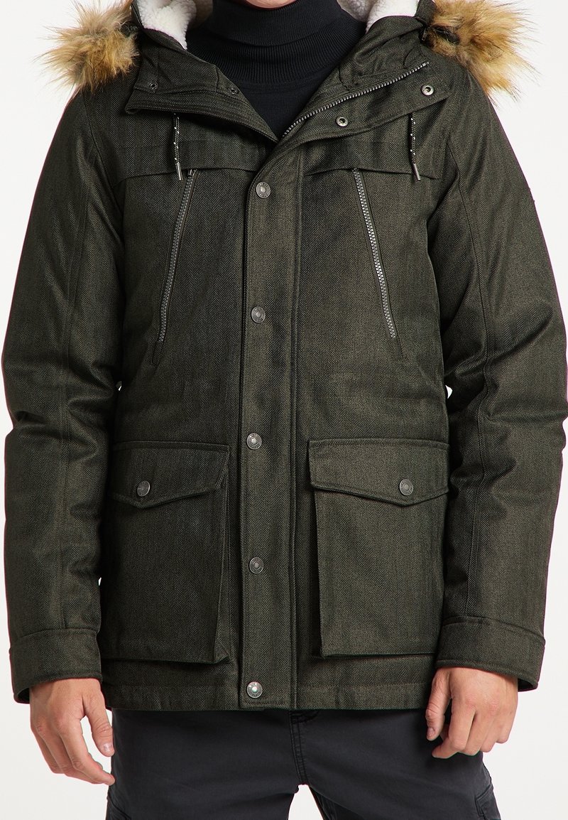 Homme portant une parka d'hiver vert foncé avec capuche bordée de fourrure, plusieurs poches zippées et à boutons, sur un pull à col roulé noir.