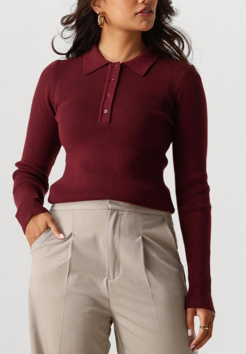 Vrouw in een bordeauxrode geribbelde lange mouwen polo trui en beige hoge taille geplooide broek, met gouden creolen en ringen.