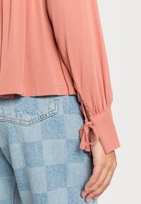 Blusa de cor pêssego com mangas longas, com punhos atados e pregas, combinada com jeans xadrez azul claro. Textura macia observada.