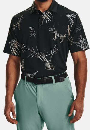 Hombre con polo negro con patrón abstracto de ramas blancas, metido en pantalones verde claro con cinturón negro, brazos relajados a los lados.