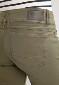 Olivgröna denimjeans med en texturerad yta, som har en bakficka och en svart läderlapp med präglad logotyp vid midjan.