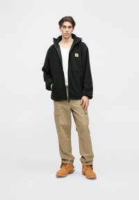 Giacca in pile nera con zip frontale, cappuccio e logo Carhartt sul lato sinistro superiore. I pantaloni cargo beige e gli stivali da lavoro arancioni completano il look.
