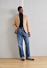 Blazer in corda a coste sottili sopra un dolcevita nero, jeans blu e scarpe eleganti multicolori. La texture liscia e la vestibilità sartoriale sono notevoli.