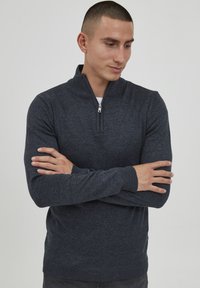 Marineblå genser med halv glidelås, ribbestrikkede ermer og kant. Laget av myk strikket stoff; har en liten logo på siden.