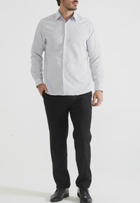 Homme portant une chemise blanche à motifs à manches longues, un pantalon noir et des chaussures de ville noires, se tenant avec une main dans sa poche sur un fond blanc uni.