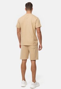 Mand set bagfra iført beige kortærmet t-shirt og matchende shorts med en baglomme, hvide sneakers, stående på hvid baggrund.