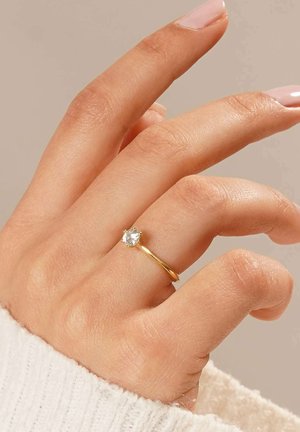 Goldring mit einem runden, klaren Edelstein, der in einer sechszackigen Fassung sitzt, getragen an einer Hand mit heller Haut. Die Hand ist teilweise mit einem weißen Stoffhandschuh bedeckt.