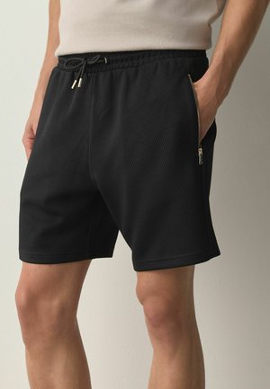 Mand iført sorte teksturerede shorts med elastisk talje og lynlås sidelommer, hænder i lommerne, beige skjorte delvist synlig.