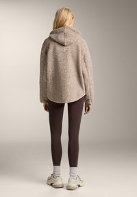 Puchaty beżowy hoodie o luźnym kroju i dużym kapturze, zestawiony z ciemno brązowymi legginsami i jasnymi sneakersami o teksturowanej podeszwie.