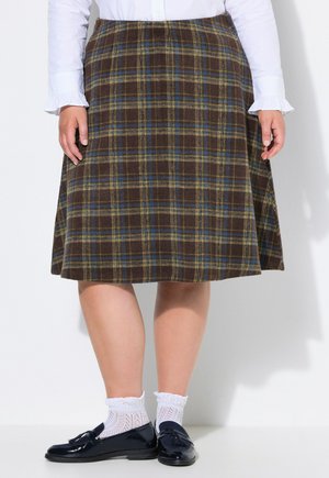 Person iført et brunt ternet nederdel til knæet, hvid skjorte med flæsemanchetter, hvide blonder sokker og sorte tassel loafers.