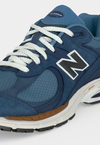 Μπλε αθλητικό παπούτσι New Balance με άσπρα κορδόνια, επάνω μέρος από δίχτυ και σουέντ, μαύρο λογότυπο "N" και λευκή σόλα με καφέ και μαύρες λεπτομέρειες.