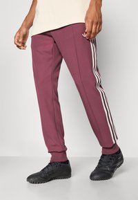 Bordowe spodnie sportowe wykonane z teksturowanej tkaniny, z trzema białymi bocznymi paskami i ściągaczami na nogawkach, w połączeniu z czarnymi korkami.