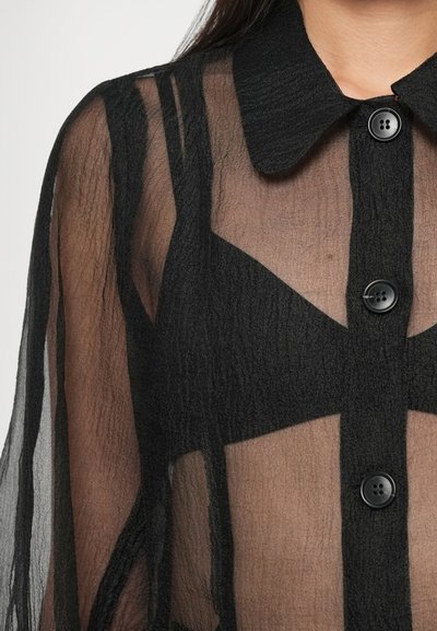 Čierna priesvitná košeľa s textúrovaným povrchom, vybavená golierom, gombíkovým zapínaním a priesvitnými panelmi, ktoré odhaľujú spodnú vrstvu oblečenia.