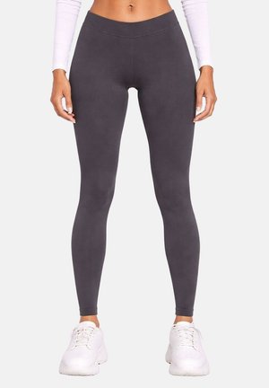 Femme portant des leggings gris foncé taille haute, un haut blanc à manches longues et des baskets blanches, debout avec les bras détendus le long du corps.