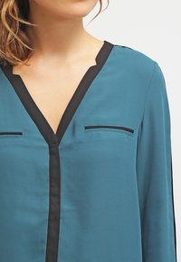 Tealblus med svart kant, V-ringning och två bröstfickor. Slätt tyg med långa ärmar och en avslappnad passform.