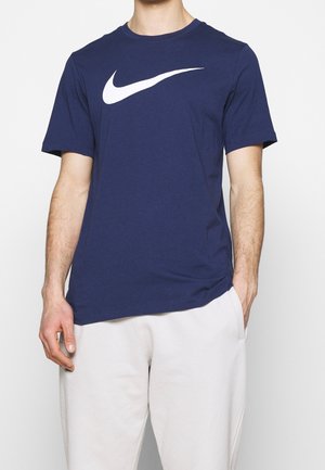 Mann, der ein navyblaues Nike-T-Shirt mit weißem Logo und hellgraue Sweatpants trägt, eine Hand in der Tasche und die andere entspannt an der Seite.