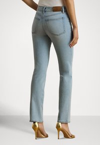 Sieviete, tērpusies gaiši zilās taisna piegriezuma džinsās un zeltainās augstpapēžu sandales, stāvot pret vienkāršu baltu fonu.