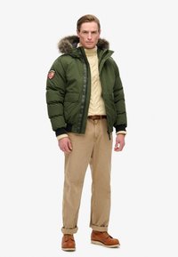 Grøn puffjakke med pelsforet hætte, sort kant og et mærke på venstre ærme. Bæres over en cremefarvet sweater og suppleret med beige bukser og brune støvler.