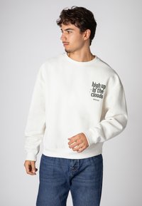Weißes Sweatshirt aus weichem Stoff, mit rundem Halsausschnitt und langen Ärmeln, mit schwarzem Text "hoch oben in den Wolken" auf der Vorderseite bedruckt.