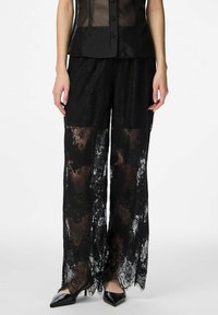 Czarne spodnie o szerokich nogawkach z koronki, charakteryzujące się półprzezroczystym materiałem, misternymi kwiatowymi wzorami oraz frędzlami na krawędziach, zestawione z czarnymi szpiczastymi szpilkami.