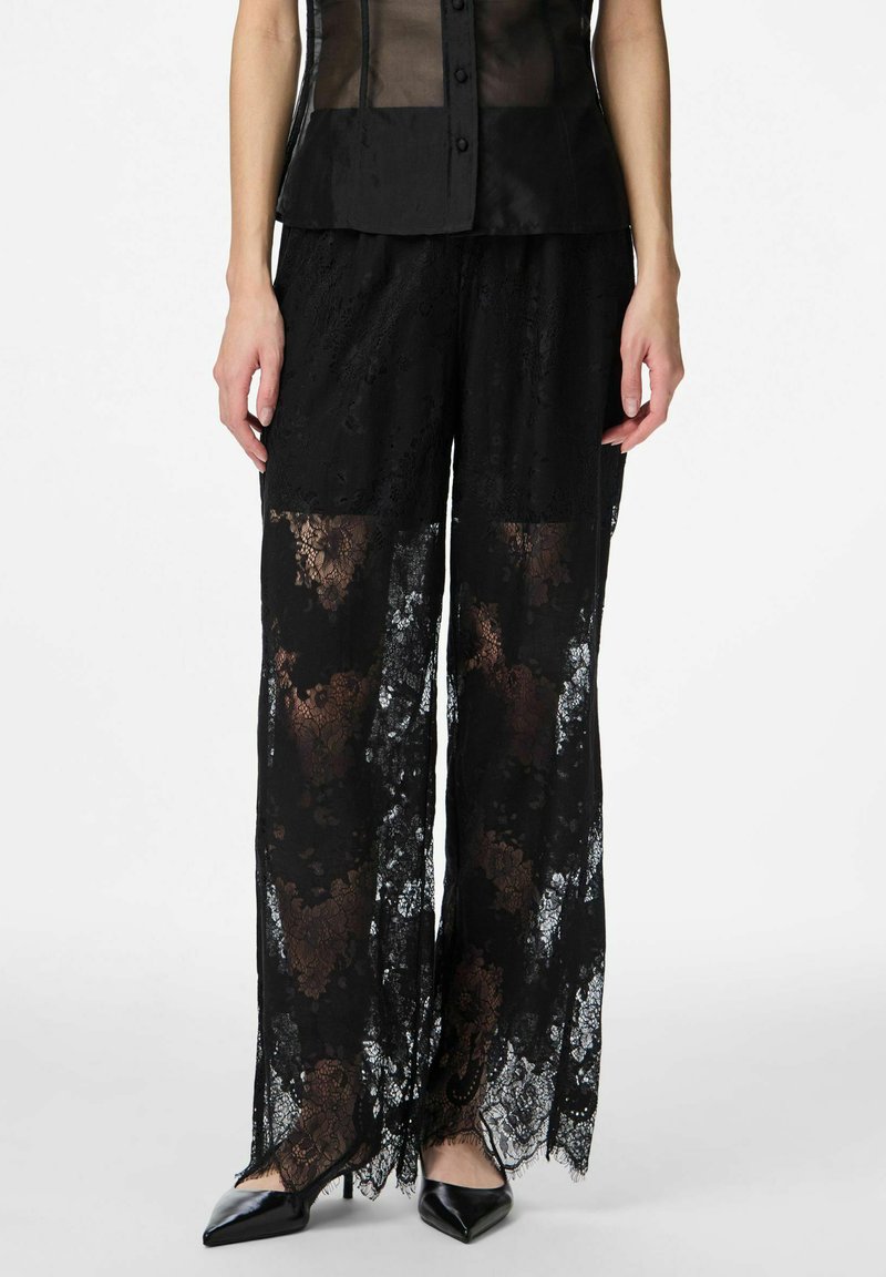 Czarne spodnie o szerokich nogawkach z koronki, charakteryzujące się półprzezroczystym materiałem, misternymi kwiatowymi wzorami oraz frędzlami na krawędziach, zestawione z czarnymi szpiczastymi szpilkami.