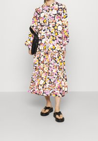 Robe à imprimé floral avec manches longues, devant à boutons, jupe fluide, fond noir avec des fleurs roses, jaunes et blanches, associée à des sandales noires.